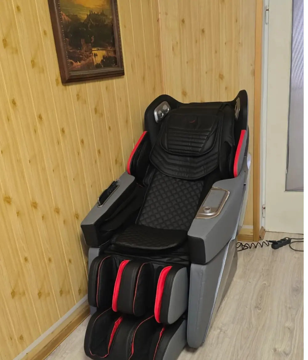 Fotel masujący Casada w czarno-szarej kolorystyce z czerwonymi akcentami, ustawiony w pomieszczeniu z drewnianą boazerią, z widocznym panelem sterowania i rozbudowanym modułem masażu nóg.