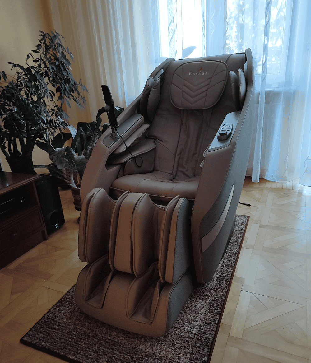 Nowoczesny fotel masujący w brązowej kolorystyce, stojący na niewielkim dywaniku w salonie, przed oknem balkonowym