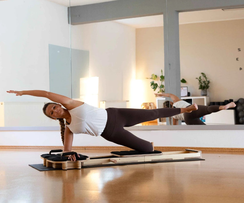 Kobieta wykonująca ćwiczenie pilates w podporze bokiem na drewnianym reformerze, z jedną nogą uniesioną i ręką wyciągniętą nad głową w jasnym studiu.
