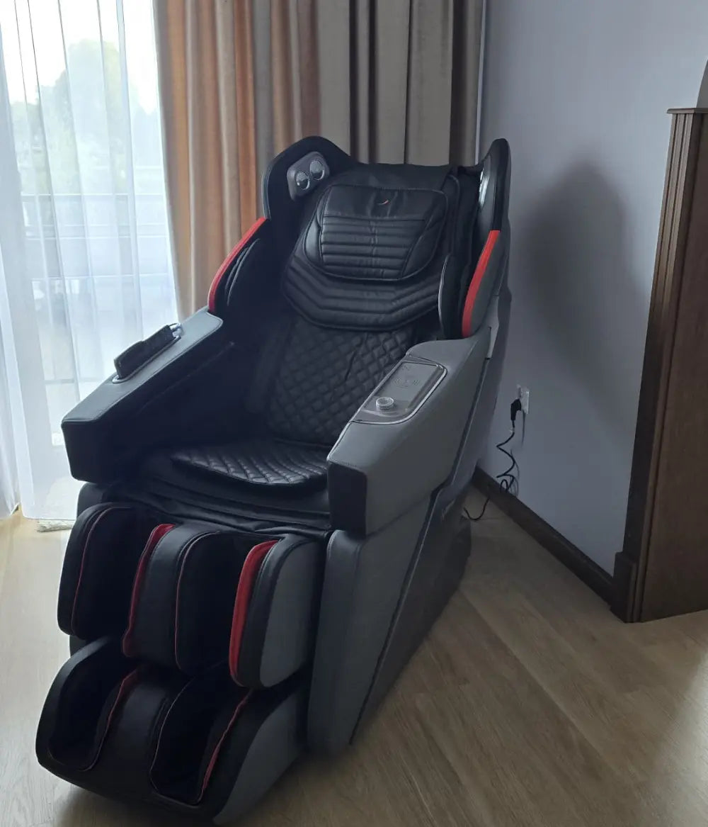 Czarno-szary fotel masujący Casada z czerwonymi akcentami, wyposażony w panel sterowania na podłokietniku i rozbudowany masaż nóg, ustawiony przy oknie z zasłonami w nowoczesnym wnętrzu.