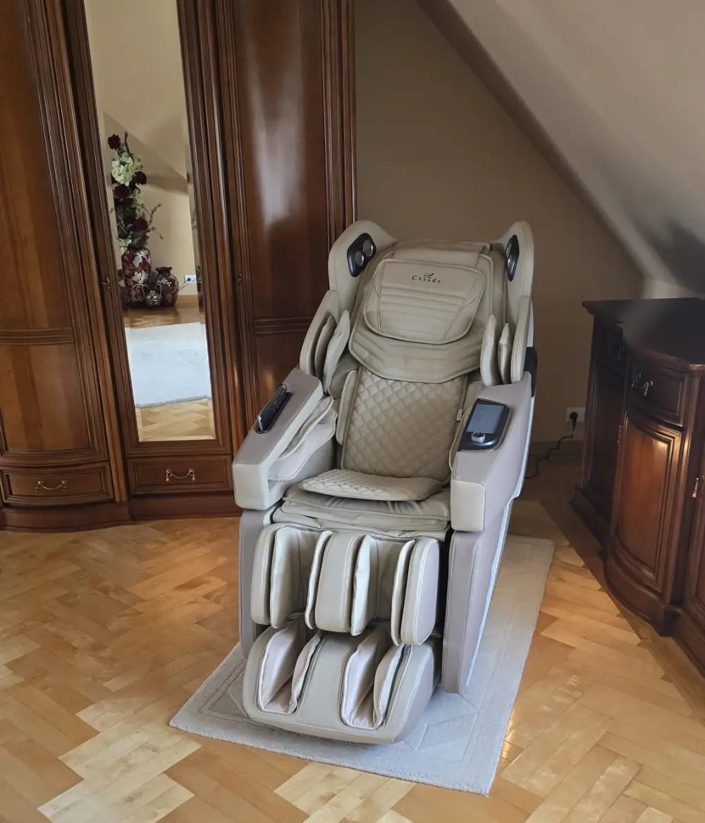 Jasny fotel masujący Casada z funkcją masażu nóg, ramion i pleców, ustawiony na jasnej macie na parkiecie, umieszczony w eleganckim wnętrzu z drewnianą zabudową i lustrem.
