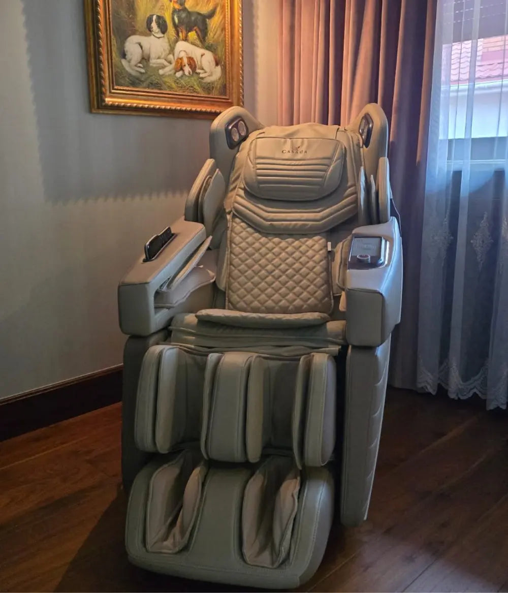 Fotel masujący Casada w jasnej tapicerce, ustawiony w eleganckim salonie obok okna z zasłonami, z widocznym panelem sterowania i rozbudowanym modułem masażu nóg.