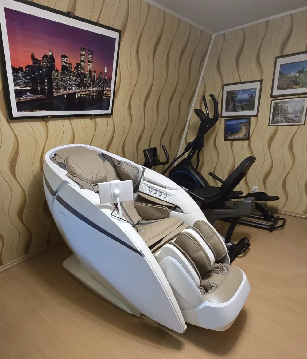 Nowoczesny fotel masujący w jasnej kolorystyce z beżowym wnętrzem znajduje się w przytulnym pokoju rekreacyjnym. Ustawiony jest na tle dekoracyjnej tapety, obok sprzętu fitness, a całość dopełniają obrazy na ścianach.